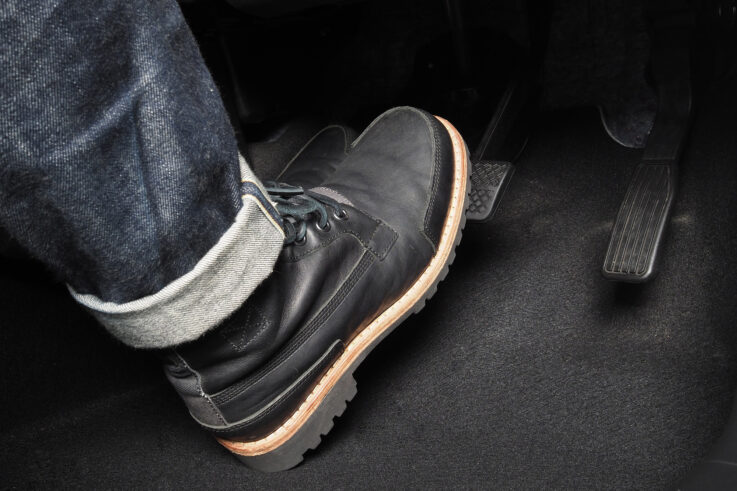 A person wearing black leather boots is pressing down on a car pedal, captured from the interior of the vehicle. The image shows the pedals and the foot in close detail, emphasizing driving actions.