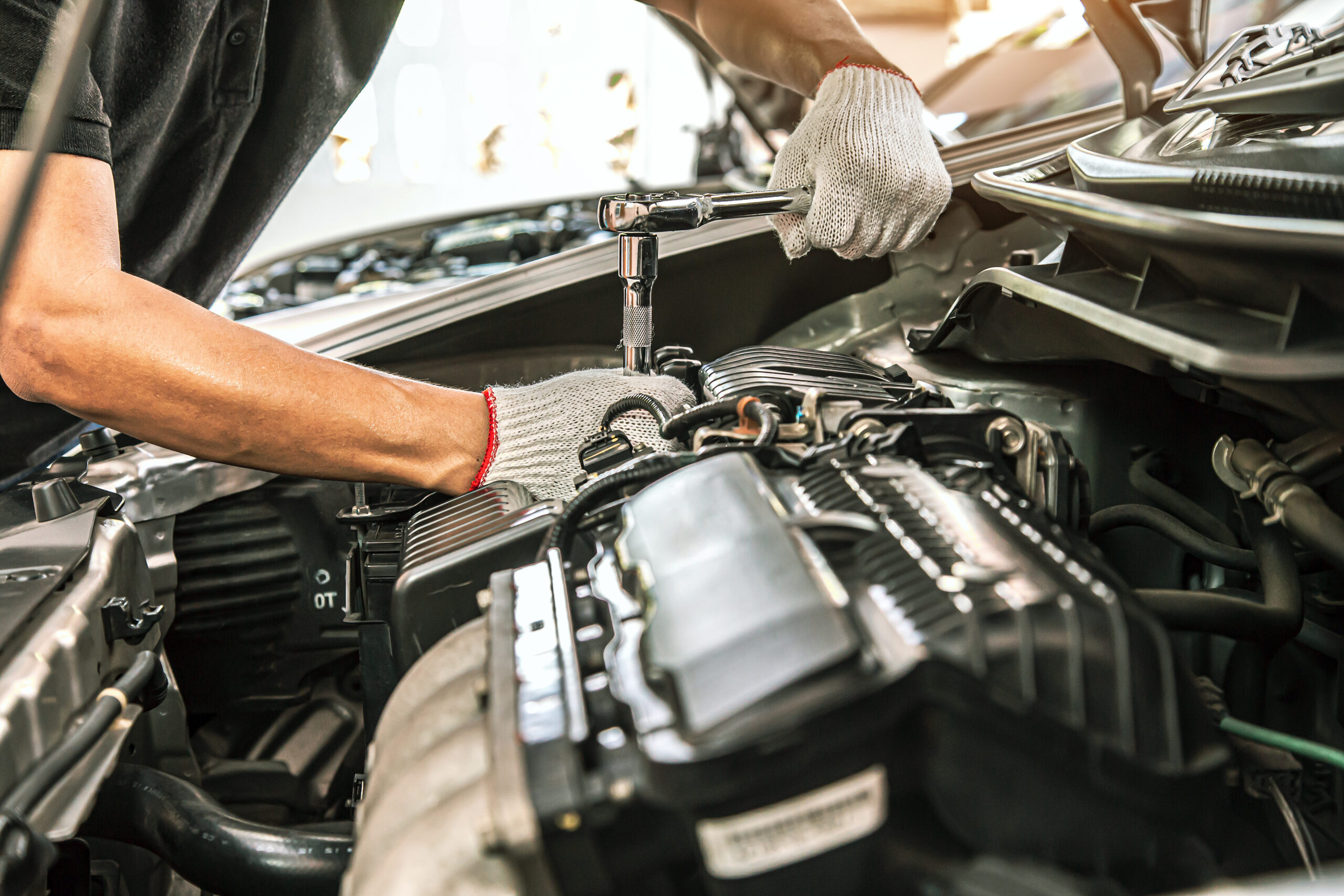 Mechanic using a socket wrench to work on a car engine, wearing protective gloves for safety while conducting repairs under the hood.