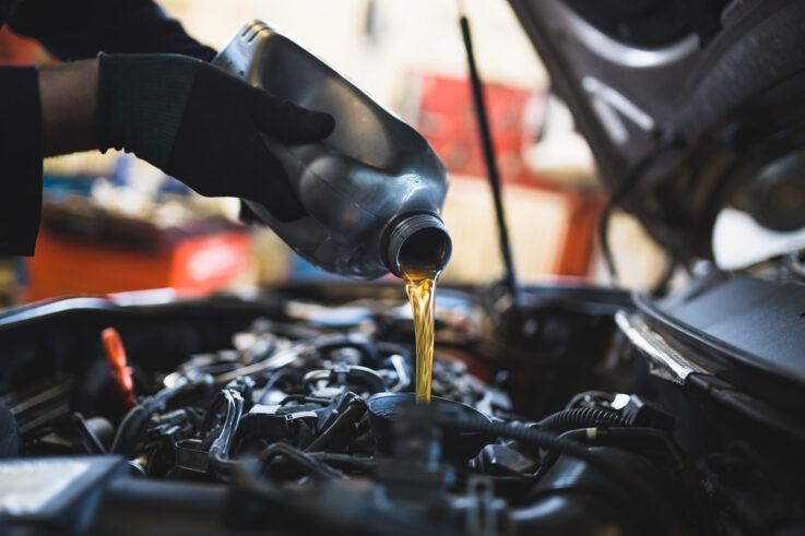 A mechanic pours fresh engine oil into a car engine with the hood open in a workshop, focusing on the oil stream and engine components.