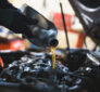 A mechanic pours fresh engine oil into a car engine with the hood open in a workshop, focusing on the oil stream and engine components.